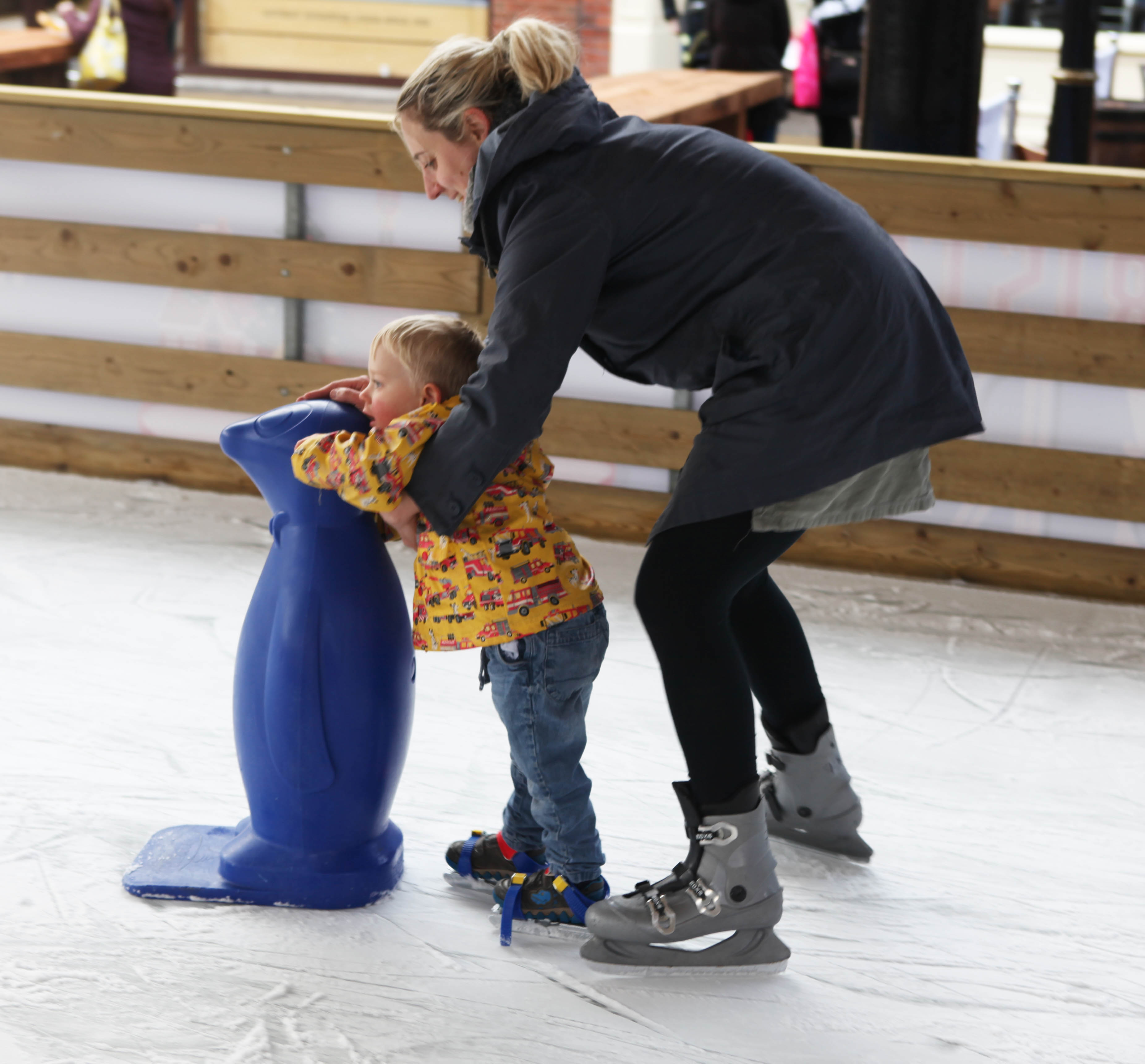 A child wearing clip over ice skates and using a penguin skate aid and help from an adult to skate. 