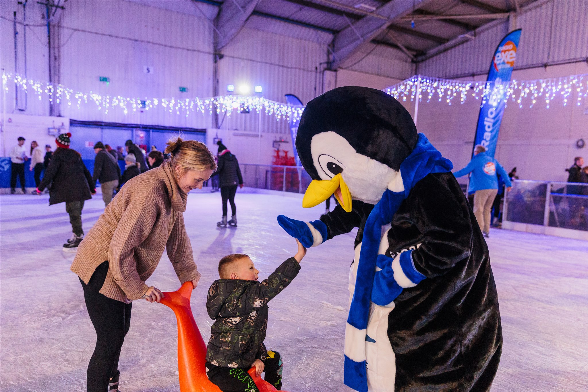 A penguin mascot high fiving a child on a seal skate aid on a winter real ice rink. 