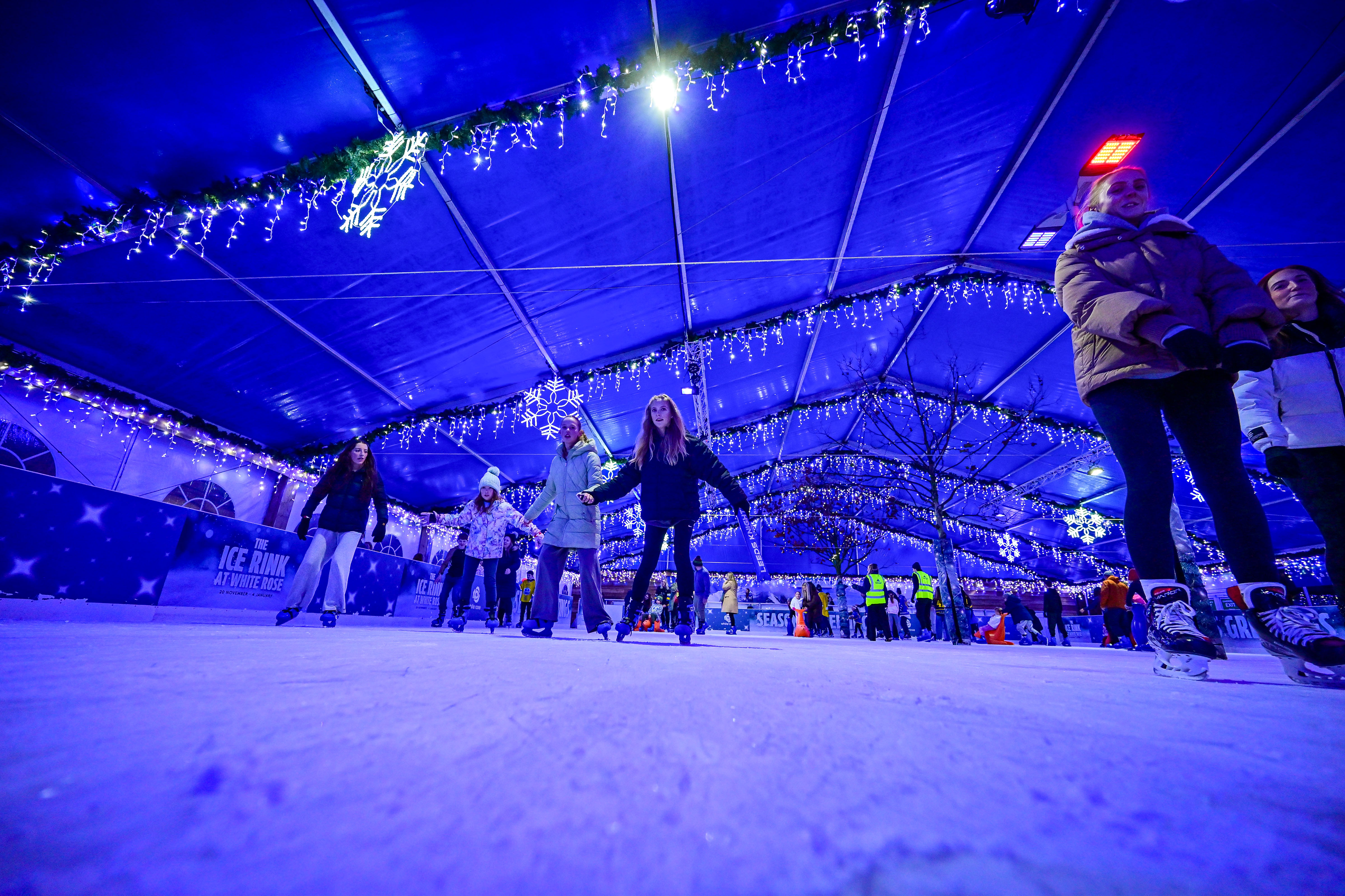 A low shot of a winter ice rink with skaters enjoying the ice. The rink is covered by a blue roofed marquee and festive lights. 