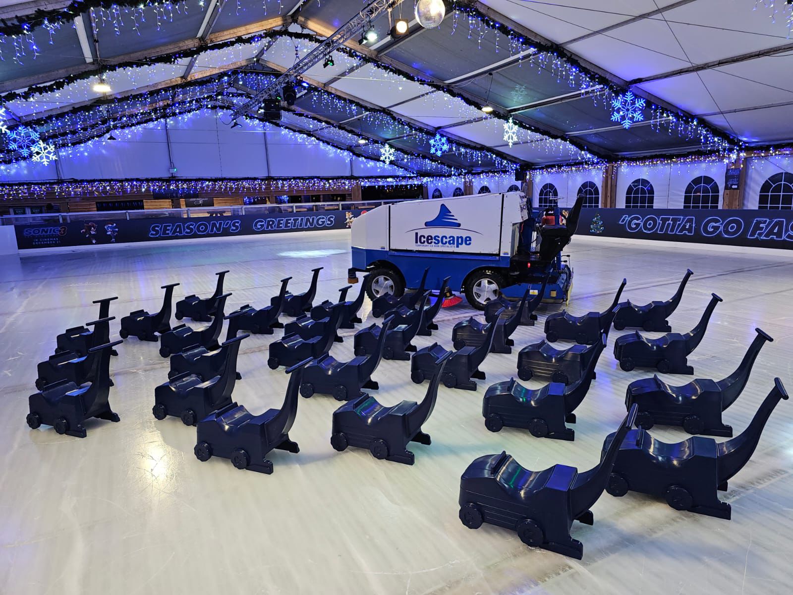 A group of Olympia Ice Resurfacer Skate aids on the ice at a winter ice rink, with a real Olympia ice resurfacer in the background. 