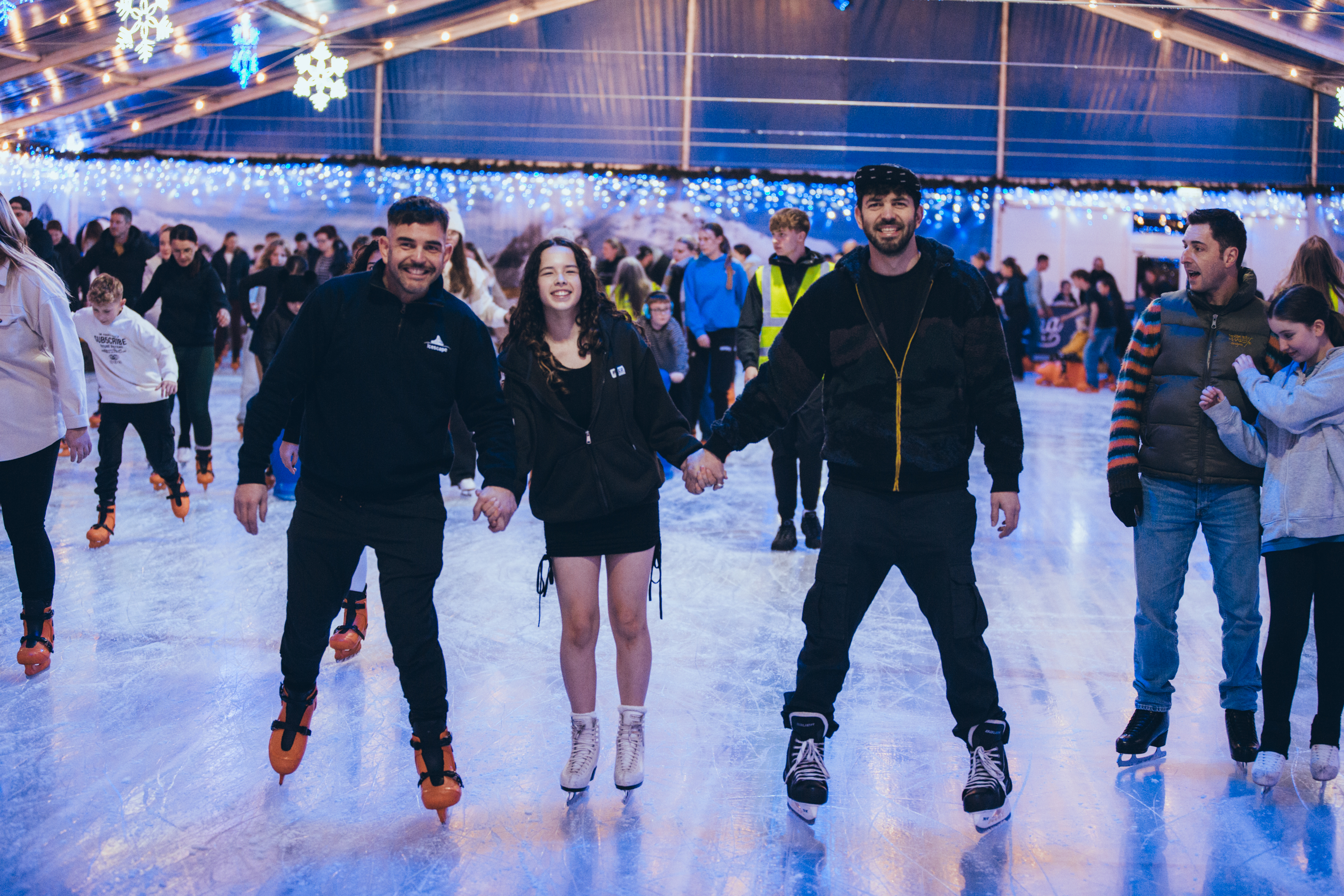 Three ice skaters holding hands on the ice and all smiling having fun at a festive ice rink. 