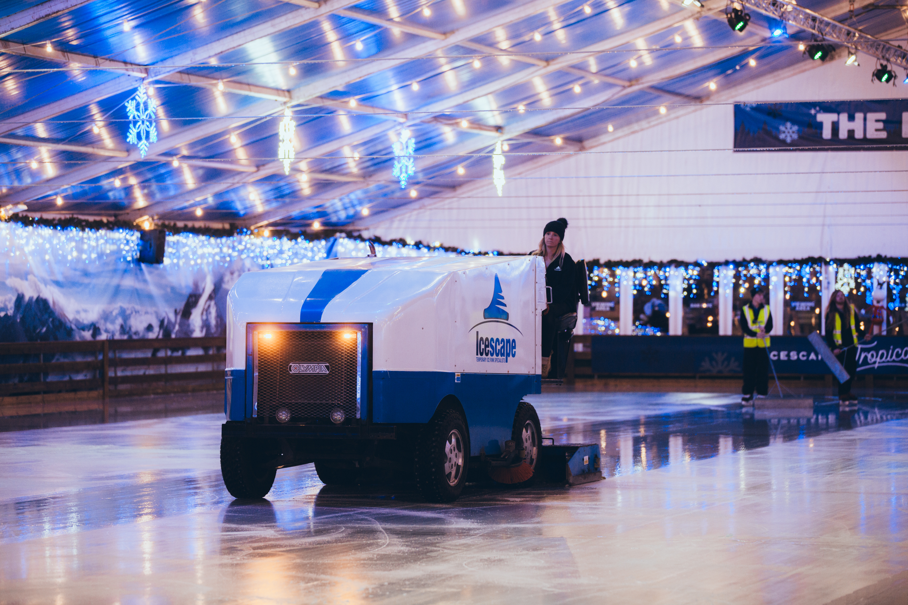 Front shot of the Olympia model ice resurfacer machine, maintaining the ice on a temporary real ice rink. Water can be seen being laid onto the ice and the machines front lights flashing. 