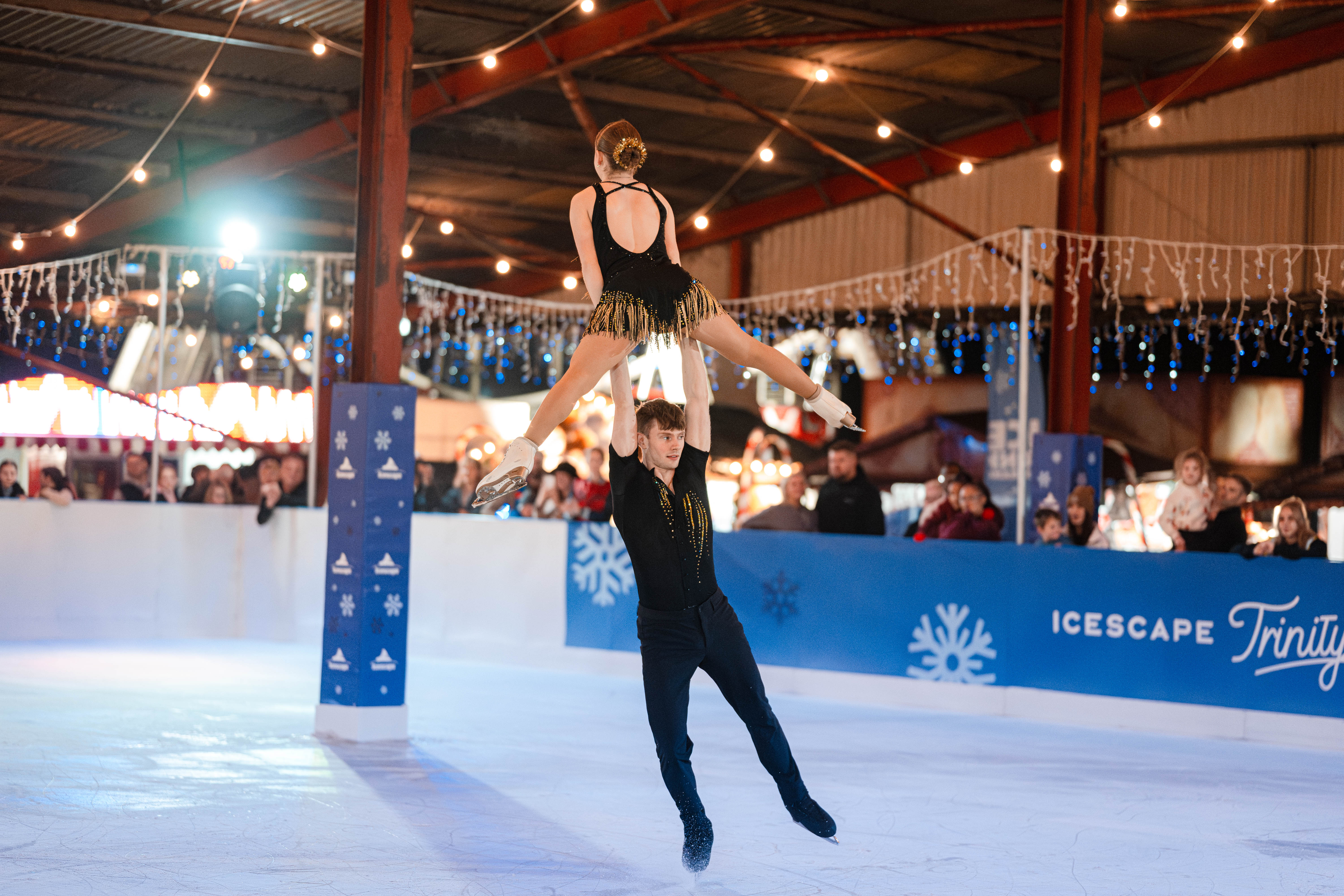 A pair of pro skaters performing a lift on a real temporary winter ice rink. In the background, the crowd is watching and enjoying the performance around the rink barrier. 